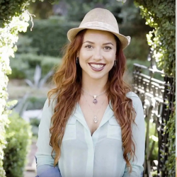 A woman with long, curly, brown hair, wearing a brown hat and white top